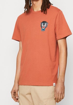 Man draagt een roest-oranje T-shirt met korte mouwen met een kleine zwarte panterafbeelding en beige cargoshorts, staand tegen een effen achtergrond.