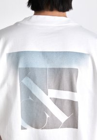 Bílá bavlněná trička s geometrickým grafickým potiskem v světle modrých a šedých tónech na zadní straně, s různými texturami a neprůhledností.