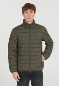 Veste matelassée vert olive avec col haut, design matelassé horizontal et fermeture zippée. Dispose de poches latérales et de poignets élastiques.