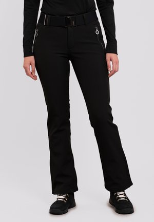 Vrouw draagt zwarte wijde broek met ritssluitingen in de zakken, zwarte riem, zwart langemouwen shirt en zwarte wandelschoenen met witte veters.