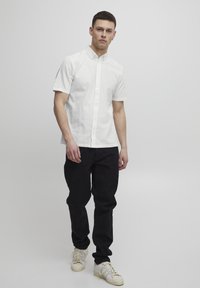 Weißes kurzärmliges Hemd mit Knopfverschluss und spitzem Kragen, kombiniert mit schwarzen Hosen und weißen Sneakern. Einfaches Design und eine klare Farbpalette.
