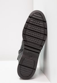 Sort sneakers med en tekstureret gummisål, der har vandrette riller og ventilationshuller. Brandlogoet er synligt på sålen.