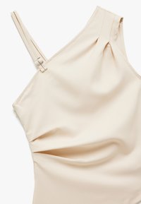 Beige one-shoulder top gemaakt van gladde stof, met een subtiele plooidetail en een verstelbare band met een zilverkleurige sluiting.