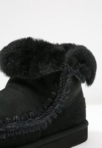 Stivale in suede nero con un morbido polsino in pelliccia, finiture decorative intrecciate e suola piatta. La superficie tessurata aumenta calore e comfort.