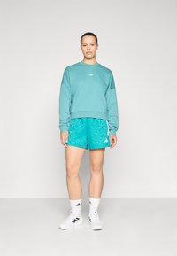 Turkos sweatshirt med rund halsringning och vit logotyp, matchad med mönstrade teal shorts och vita träningsskor samt strumpor.