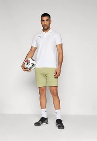 Mees valges polos ja beežides lühikestes pükstes hoiab valget ja musta jalgpalli, kannab valgeid sokke ja musti krampe, seisab halli taustaga taustal.