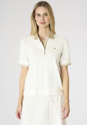 Donna con lunghi capelli biondi che indossa una polo bianca a maniche corte con colletto beige e gonna bianca, in piedi davanti a uno sfondo semplice.