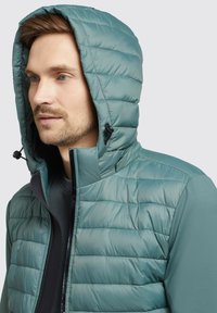 Gequiltete Jacke in Teal mit Kapuze. Verfügt über einen verstellbaren Kordelzug am Hals und kontrastierendes Material an den Seiten.