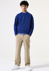 Sudadera azul con puños acanalados, superpuesta a una camisa de cuadros. Pantalones beiges con bolsillos de parche, combinados con zapatillas blancas.