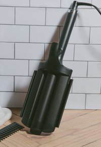 Outil de coiffure noir avec un baril large et plat et trois sections arrondies. Dispose d'un long manche et d'un cordon électrique, sur fond de carreaux blancs.
