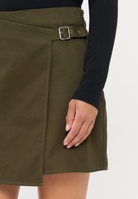 Olijfgroene rok tot de knie van katoen, met een zijgesp detail, een lichte A-lijn vorm en zichtbare stiksels langs de zoom.