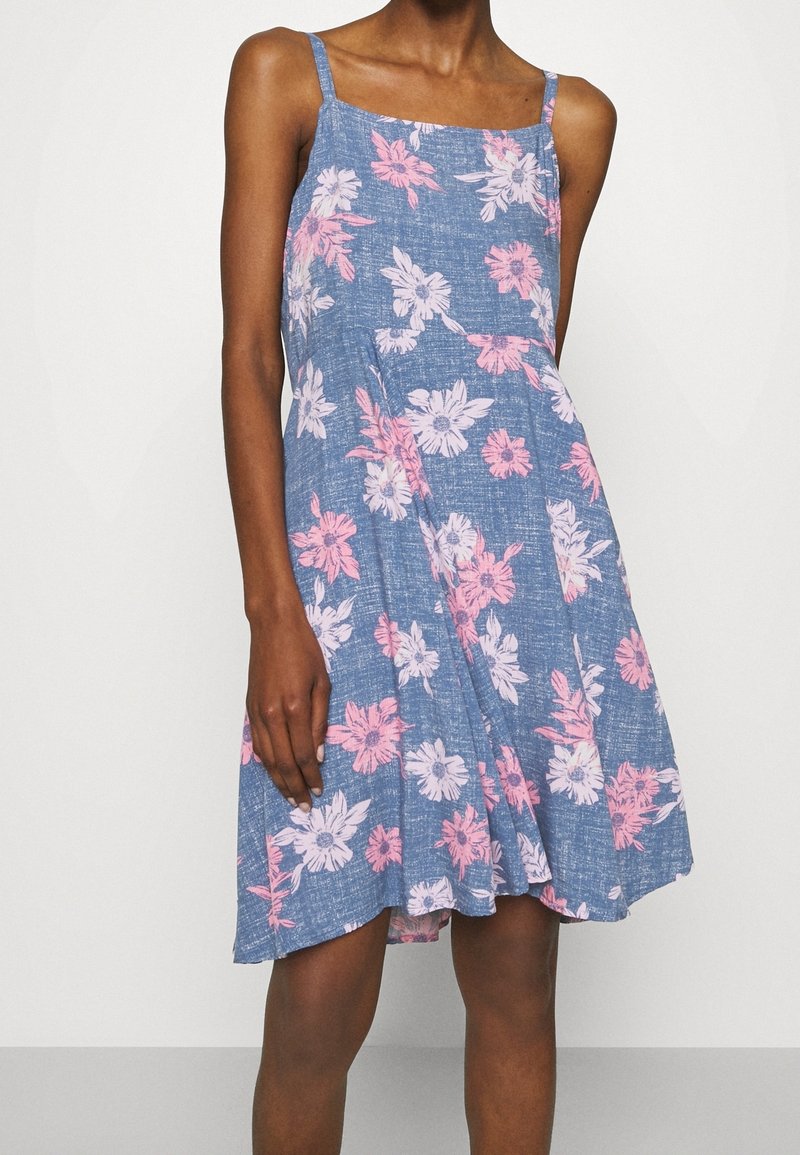 Femme portant une robe sans manches bleue à fleurs roses et blanches, debout devant un fond blanc uni.