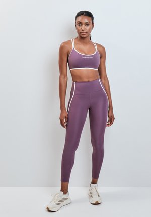 Vrouw staat tegen een muur, draagt een paarse sportbeha en bijpassende hoog waist legging met witte sneakers.
