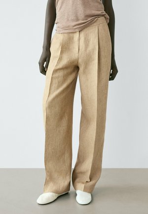 Personne portant un pantalon en lin beige à taille haute et jambes larges, une chemise marron clair rentrée, et des chaussures blanches, se tenant sur un sol uni.