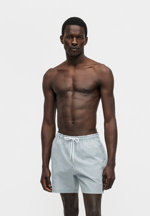 Slim dark-skinned man with short hair wearing light blue and white striped swim shorts with drawstring, standing against plain gray background.