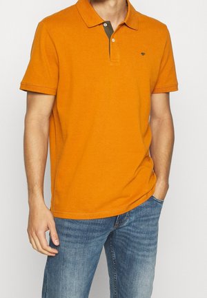Homme portant un polo orange à manches courtes avec une patte de boutonnage sombre en contraste et un jean bleu, main dans la poche.