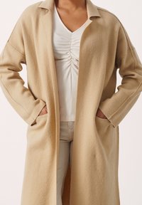 Beige wollen lange jas met een kraaghalslijn, losse pasvorm, zijzakken en een zachte textuur. Draag het over een witte geraffineerde top en lichte broek.
