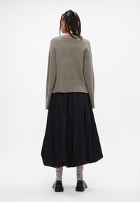 Oliwkowy sweter z dzianiny o luźnym kroju, zestawiony z czarną spódnicą do kostek i szarymi skarpetkami w paski, noszony z czarnymi butami.