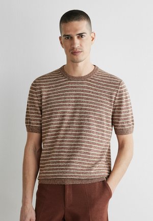 Mand med kort hår iført brun og beige stribet kortærmet skjorte og brune bukser, stående mod en ensfarvet lys væg.