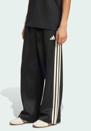 Osoba ubrana w luźne czarne spodnie dresowe Adidas z trzema białymi paskami po bokach oraz białe sportowe buty.