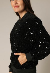 Veste bomber noire à sequins avec une surface texturée, des poignets et un ourlet côtelés, ainsi que des poches latérales. Des détails scintillants rehaussent son design.