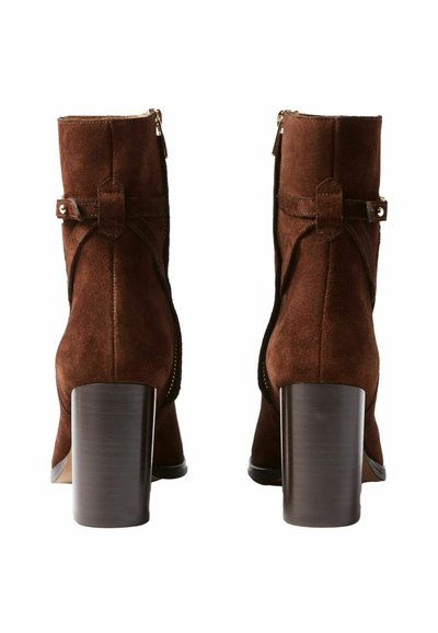 Botines de ante marrón con tacón grueso, que cuentan con una cremallera lateral y una correa decorativa con detalles metálicos.