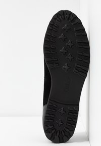 Semelle de chaussure noire avec des empreintes profondes et des motifs en forme de croix, inclinée contre un fond blanc aux coins.