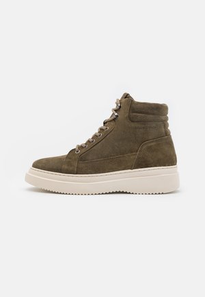 Oliwkowe zamszowe trampki typu high-top z kremową gumową podeszwą, z metalowymi oczkami i beżowymi sznurowadłami. Szczegół z logo po boku.