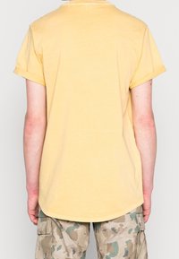 Yellow cotton t-shirt with rolled sleeves, curved hem, and a soft texture, paired with camouflage cargo pants featuring pockets.