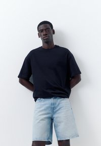 Jeune homme debout contre un mur blanc, portant un t-shirt noir oversize et un short en jean bleu clair, mains derrière le dos, expression neutre.