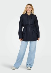 Veste à capuche bleu marine au design court, taille ajustable avec cordon de serrage, et deux poches frontales ; associée à un jean large bleu clair et des baskets.