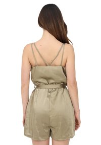 Mujer con el cabello largo y marrón vestida con un mono sin mangas de satén beige, con tirantes traseros finos en forma de cruz y un lazo en la cintura, de espaldas.