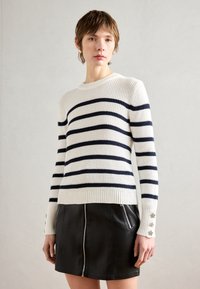 Hvitstripet genser med marineblå horisontale striper, ribbestrikket trim og stjernedetaljer på ermene, sammen med et svart skinnskjørt med glidelås.