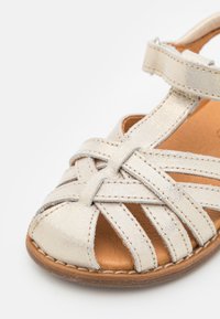Sandalia de cuero plateado con tiras trenzadas, diseño de punta abierta y plantilla de cuero marrón. Presenta detalles de costura y correa ajustable en el tobillo.