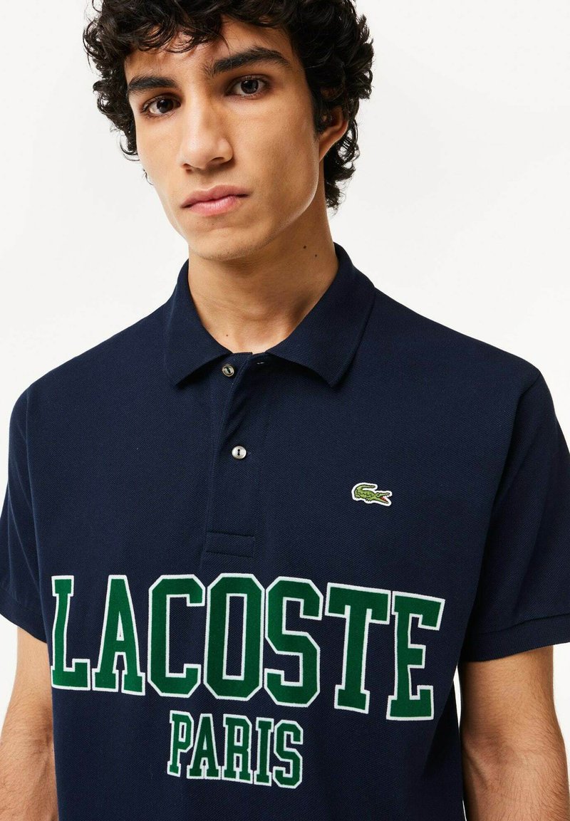 Polo marine avec le lettrage vert "LACOSTE PARIS" et un petit logo crocodile. Il présente un col classique et une patte de boutonnage à deux boutons.