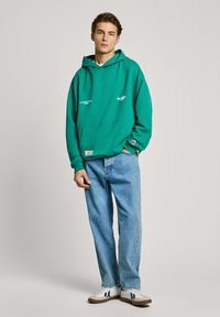 Sudadera con capucha verde y un bolsillo frontal, combinada con pantalones vaqueros sueltos de color azul claro y zapatillas blancas. Presenta costuras decorativas y parches.