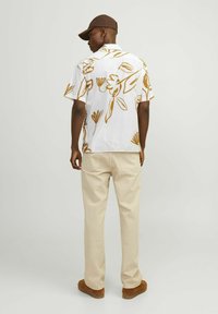 Camicia bianca a maniche corte con motivi floreali marroni, abbinata a pantaloni beige e scarpe marroni. Persona con un cappello marrone.
