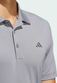 Harmaa polo-paita, jossa on kaulus, kaksi nappia ja lyhyet hihat. Vasemmassa rinnassa on tummanharmaa kolmikulmainen logo. Sujuva, tekstuuriainen kangas.