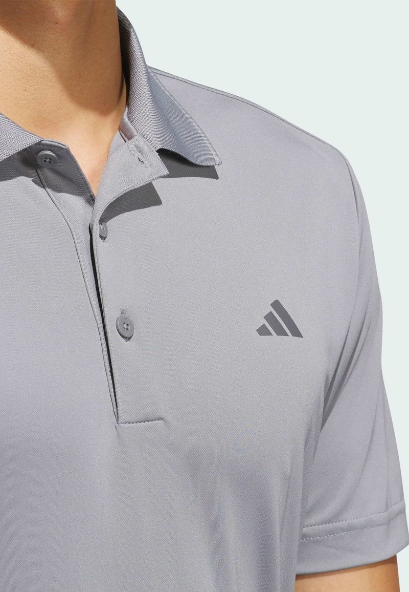 Polo gris avec col, deux boutons et manches courtes. Présente un logo triangulaire gris foncé sur la poitrine gauche. Tissu lisse et texturé.