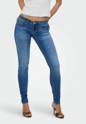 Kobieta ubrana w niebieskie, przylegające spodnie z podwyższonym stanem, biały krótki top i brązowe wysokie sandały na odkryte palce, stojąca z rękami za plecami.