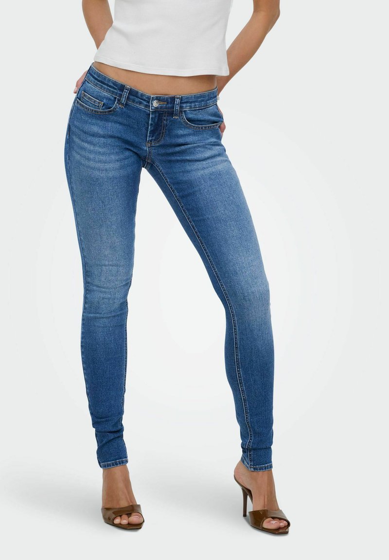 Femme portant un jean skinny bleu taille moyenne, un haut blanc court et des talons ouverts marron, debout les mains derrière le dos.