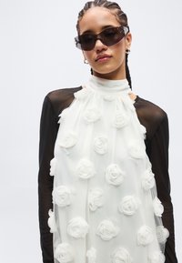 Kobieta z plecionymi włosami, nosząca ciemne okulary przeciwsłoneczne i białą sukienkę z 3D kwiatowymi aplikacjami na czarnych przezroczystych rękawach.