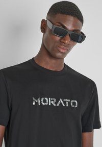 Camiseta de algodón negra con un logo texturizado y reflectante "MORATO" en el pecho. El modelo lleva unas gafas de sol negras angulares.