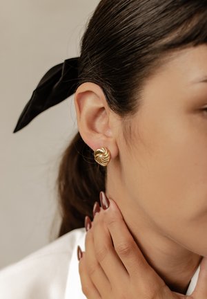 Gold spiral stud earring worn on the left ear, featuring a textured, twisted design. Neat black ribbon in hair, hand gently touching neck.