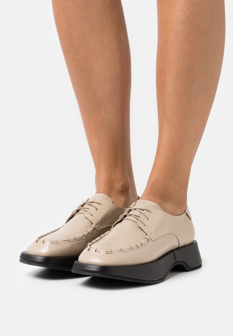Chaussures en cuir beige à lacets avec semelles épaisses noires portées pieds nus, mettant en valeur les détails de la couture et le design moderne de la chaussure.