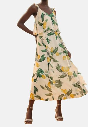 Femme portant une robe crème fluide sans manches avec un imprimé citrons jaunes et feuilles vertes, associée à des sandales beiges à talons hauts.