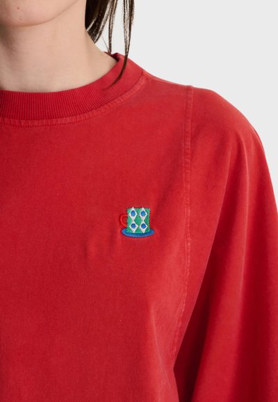 Personne portant un sweat-shirt rouge avec un petit motif brodé de tasse verte sur la poitrine près du col sur un fond uni.