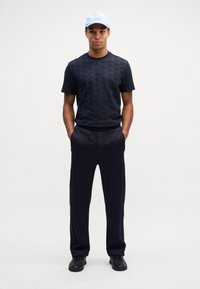 T-shirt nera con motivo, colletto a giro, pantaloni scuri e sneakers nere; completato con un cappellino azzurro chiaro con il logo.