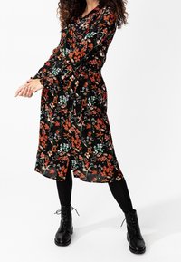 Abito con stampa floreale a maniche lunghe, sfondo nero, fiori rossi, arancioni e verdi vibranti. Indossato con stivali neri con lacci e leggings.