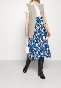 Chaleco beige sin mangas sobre una camisa blanca con cuello, combinado con una falda azul con estampado floral. Botas negras hasta la rodilla. Bolso blanco. Texturas suaves.